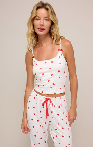 A woman wears a white pajama set with red heart prints, featuring a cropped camisole with delicate lace trim, red bows on the straps, and matching drawstring pants. The cozy, romantic loungewear exudes a playful and feminine charm.