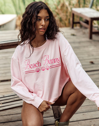 Woman wearing a pink 'Beach Bunny' sweatshirt sitting on a wooden deck.