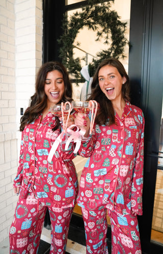 Two women wearing festive pink pajamas with holiday patterns, holding a gift bag.