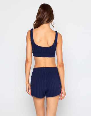 Woman wearing a navy blue ribbed knit bralette crop top with a scoop neckline and wide straps, standing against a plain white background.
Woman wearing navy blue ribbed knit lounge shorts with a high-rise drawstring waistband, standing barefoot against a plain white background.