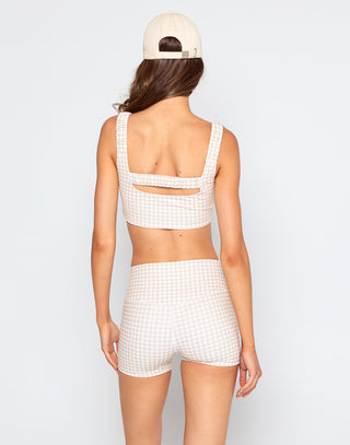 A woman wearing a beige and white gingham crop tank with wide ruffled straps and matching high-waisted gingham biker shorts, styled in a soft, neutral activewear look.