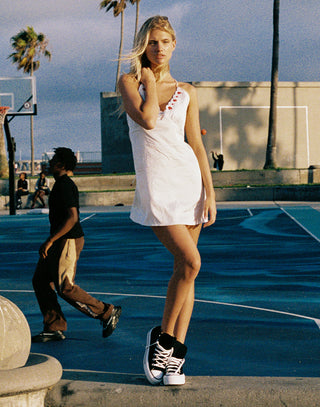 A woman stands on an outdoor basketball court wearing a white sleeveless mini dress with red lace-up details at the neckline and black-and-white high-top sneakers. Palm trees and a building are visible in the background, with another person walking nearby.